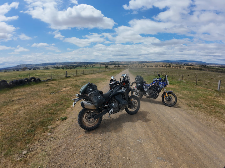 Two adventure motorcycles parked on a dirt road in an open grassy landscape under a blue sky with white clouds. One motorcycle is black with luggage and a rider in a white helmet sits on it, while another blue and yellow motorcycle stands nearby.